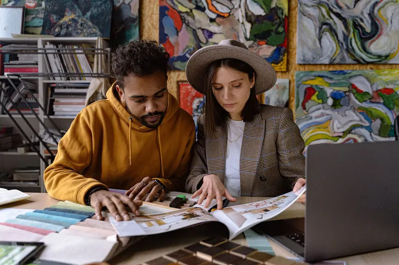Two people at a studio desk comparing colour swatches and a printed catalogue, paintings covering the wall behind them