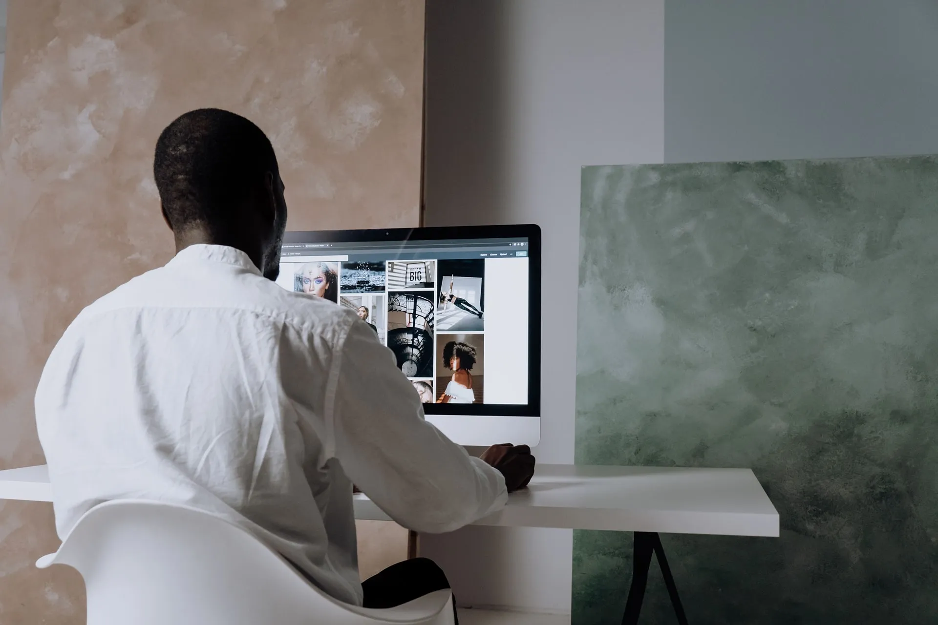 A person working at a desktop computer showing a grid of interior photographs, in a plain plastered room