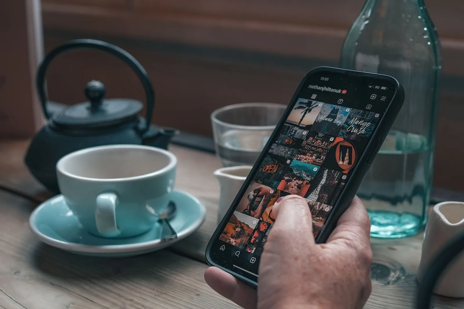A hand holding a phone showing a grid of social media photographs, beside a teapot and a cup of tea